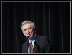 Middle-aged man presenting on a stage with dark background.